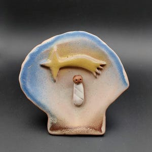Può includere: Una scena della natività in ceramica con un bambino Gesù in un pannolino bianco, una testa marrone e una stella gialla sopra. La scena è dipinta in tonalità di blu, giallo e marrone.