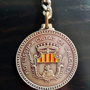 May include: A silver-toned keychain with a round medal featuring a coat of arms with red and yellow stripes, a crown, and the text "FIDELISSIMA CIUTAT DE L'ALGUER" in a circle.