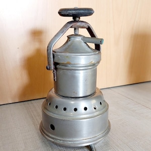 Puede incluir: Una cafetera de espresso vintage de color plateado con una perilla negra en la parte superior. La cafetera tiene una base redonda con agujeros para la distribución del calor y tres patas.