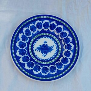 May include: A round, white ceramic plate with a blue floral design. The plate features a central floral motif and a border of blue flowers and green dots. The plate is likely used for serving food.