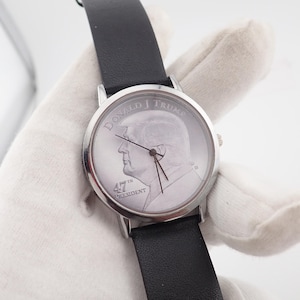 May include: A silver-toned wristwatch with a black leather band. The watch face features a profile of Donald Trump, with the text "DONALD J TRUMP" and "47TH PRESIDENT". The watch has black hands and a silver bezel.