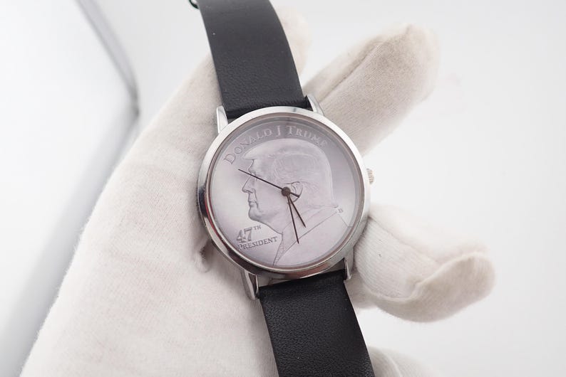 May include: A silver-toned wristwatch with a black leather band. The watch face features a profile of Donald Trump, with the text "DONALD J TRUMP" and "47TH PRESIDENT". The watch has black hands and a silver bezel.