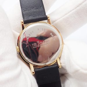 May include: Close-up of a gold-toned watch with a black leather band. The watch face is visible, with the text "Le Jour" and "Stainless Steel Back" on the back of the watch. The watch is set against a white background.