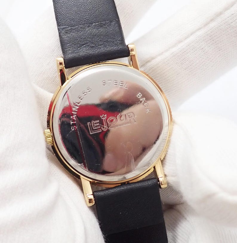 May include: Close-up of a gold-toned watch with a black leather band. The watch face is visible, with the text "Le Jour" and "Stainless Steel Back" on the back of the watch. The watch is set against a white background.