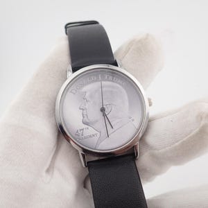 May include: Silver-toned wristwatch with a black leather band. The watch face features a profile image and the text "DONALD J TRUMP 47TH PRESIDENT". The watch has a classic design.