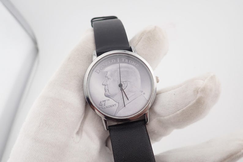 May include: Silver-toned wristwatch with a black leather band. The watch face features a profile image and the text "DONALD J TRUMP 47TH PRESIDENT". The watch has a classic design.