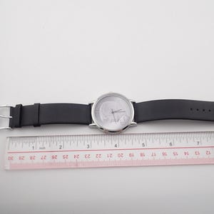 May include: A silver-toned wristwatch with a black leather band is displayed next to a ruler. The watch face is white with silver hands. The ruler shows measurements in inches.