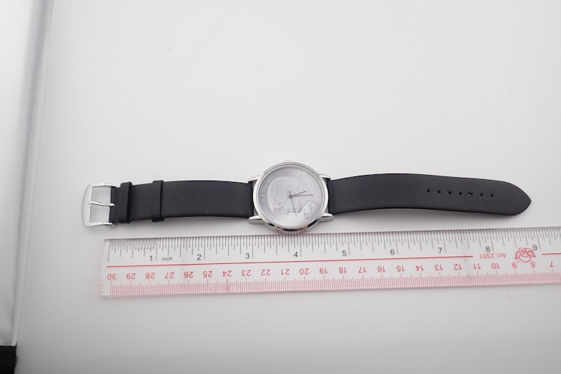 May include: A silver-toned wristwatch with a black leather band is displayed next to a ruler. The watch face is white with silver hands. The ruler shows measurements in inches.