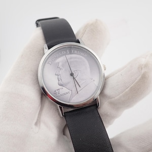 May include: A silver-toned wristwatch with a black leather band. The watch face features a profile of Donald Trump and the text "DONALD J TRUMP" and "47th PRESIDENT". The watch has a classic design.