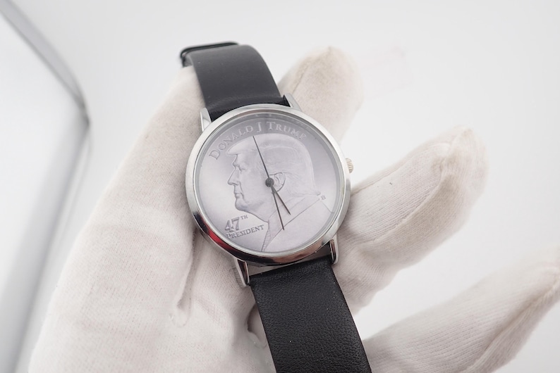 May include: A silver-toned wristwatch with a black leather band. The watch face features a profile of Donald Trump and the text "DONALD J TRUMP" and "47th PRESIDENT". The watch has a classic design.