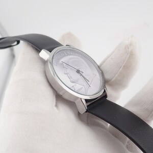 May include: A silver-toned wristwatch with a black leather band. The watch face features a portrait of a man and the words "45th PRESIDENT". The watch is held against a white background.
