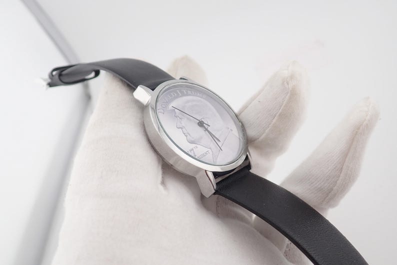 May include: A silver-toned wristwatch with a black leather band. The watch face features a portrait of a man and the words "45th PRESIDENT". The watch is held against a white background.