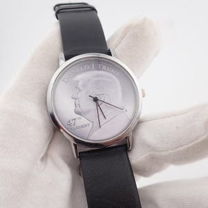 May include: Silver-toned wristwatch with a black leather band. The watch face features a profile portrait of Donald Trump and the text "DONALD J TRUMP" and "47th PRESIDENT". The watch has black hands and a silver bezel.