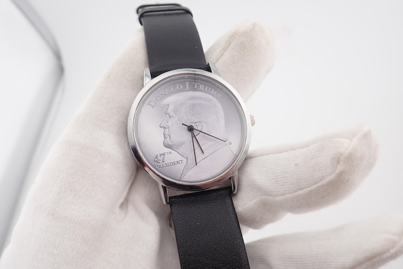 May include: Silver-toned wristwatch with a black leather band. The watch face features a profile portrait of Donald Trump and the text "DONALD J TRUMP" and "47th PRESIDENT". The watch has black hands and a silver bezel.