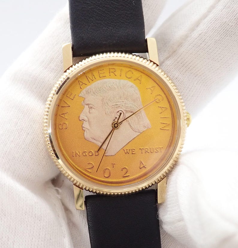 May include: Gold-toned wristwatch with a black leather band. The watch face features a gold coin design with the text "SAVE AMERICA AGAIN" and the year "2024". The watch has gold-colored hands and a gold-colored bezel.