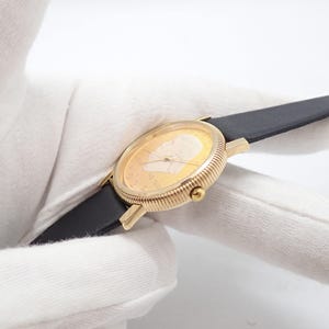 May include: A gold-toned wristwatch with a black leather band. The watch face features a detailed relief of a person's profile. The watch has a ribbed bezel and a small crown. The watch band is approximately 0.75 inches wide.