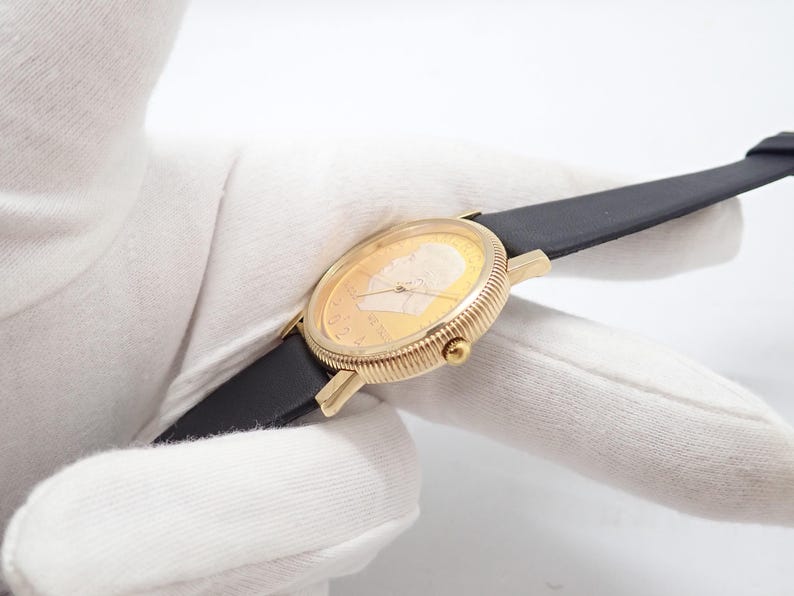 May include: A gold-toned wristwatch with a black leather band. The watch face features a detailed relief of a person's profile. The watch has a ribbed bezel and a small crown. The watch band is approximately 0.75 inches wide.