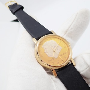 May include: A gold-toned wristwatch with a black leather band. The watch face features a portrait and the text "SAVE AMERICA AGAIN" and "IN GOD WE TRUST 2024". The watch has gold hands and a gold buckle.