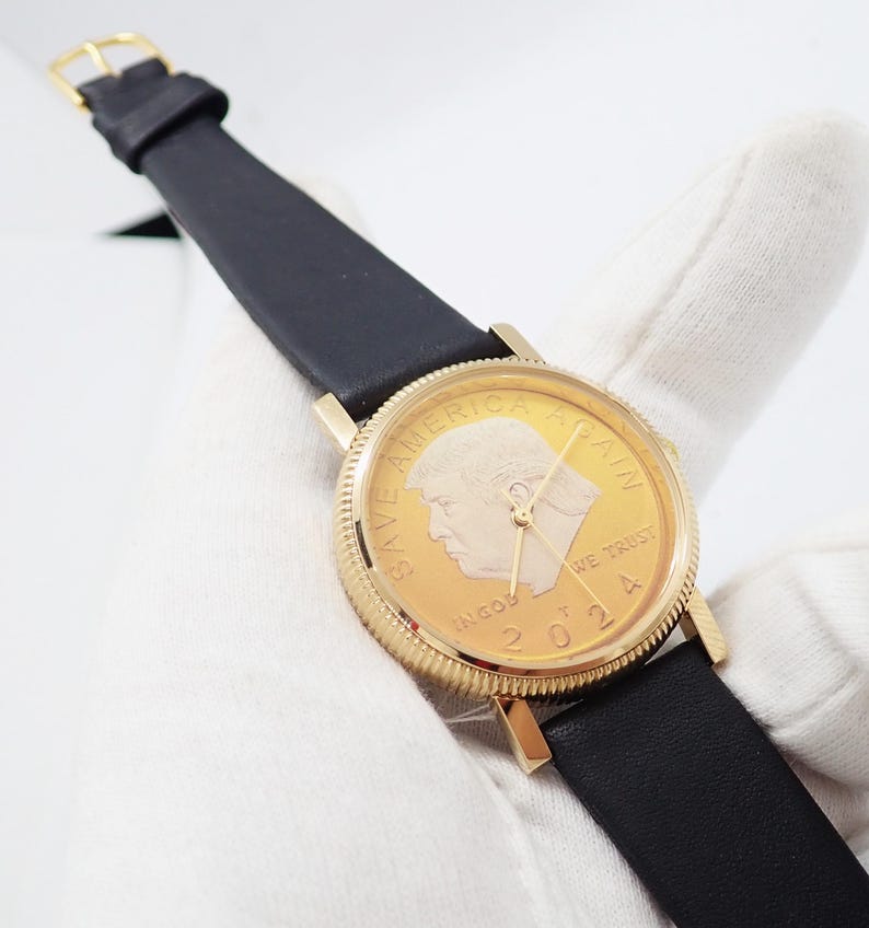 May include: A gold-toned wristwatch with a black leather band. The watch face features a portrait and the text "SAVE AMERICA AGAIN" and "IN GOD WE TRUST 2024". The watch has gold hands and a gold buckle.