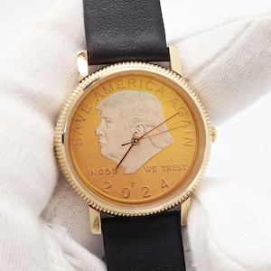 May include: A gold-toned wristwatch with a black leather band. The watch face features a portrait and the text "SAVE AMERICA AGAIN" and "IN GOD WE TRUST 2024". The watch has gold hands and a gold bezel.