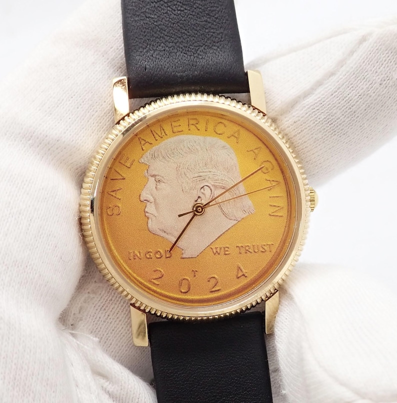 May include: A gold-toned wristwatch with a black leather band. The watch face features a portrait and the text "SAVE AMERICA AGAIN" and "IN GOD WE TRUST 2024". The watch has gold hands and a gold bezel.
