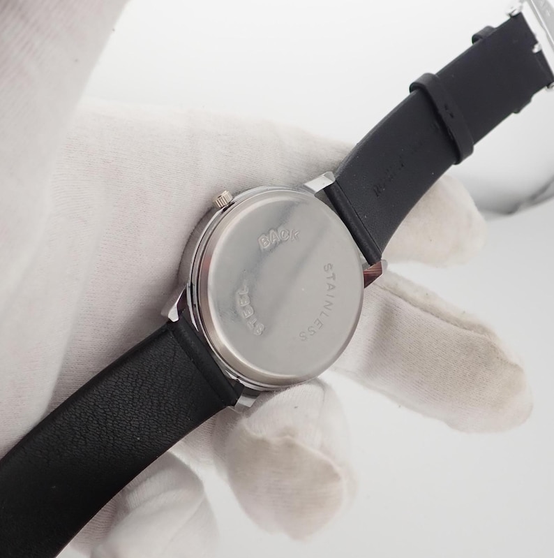 May include: A silver-toned wristwatch with a black leather band. The watch face is round with a stainless steel back. The band is attached to the watch with silver-toned metal. The watch is shown from the back.