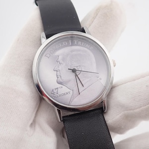 May include: A silver-toned wristwatch with a black leather band. The watch face features a profile of Donald J. Trump, with the text "DONALD J TRUMP" and "47TH PRESIDENT". The watch has black hands and a silver bezel.