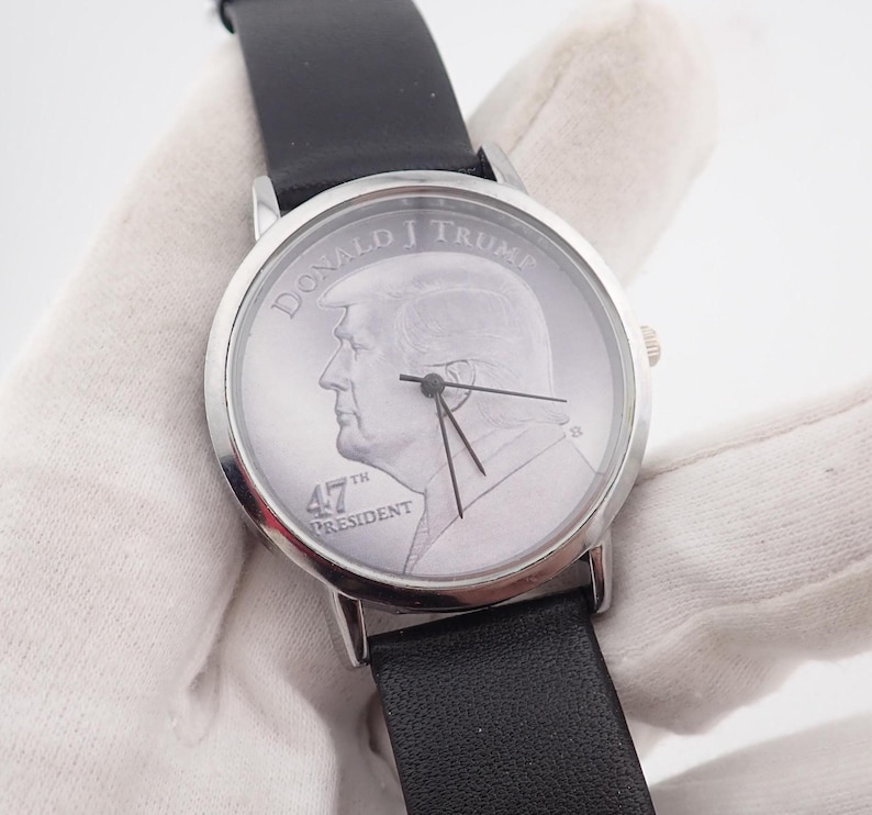 May include: A silver-toned wristwatch with a black leather band. The watch face features a profile of Donald J. Trump, with the text "DONALD J TRUMP" and "47TH PRESIDENT". The watch has black hands and a silver bezel.