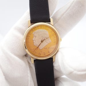 May include: Gold-toned wristwatch with a black leather band. The watch face features a profile portrait and the text "SAVE AMERICA AGAIN" and "IN GOD WE TRUST 2024". The watch has a gold-colored bezel.