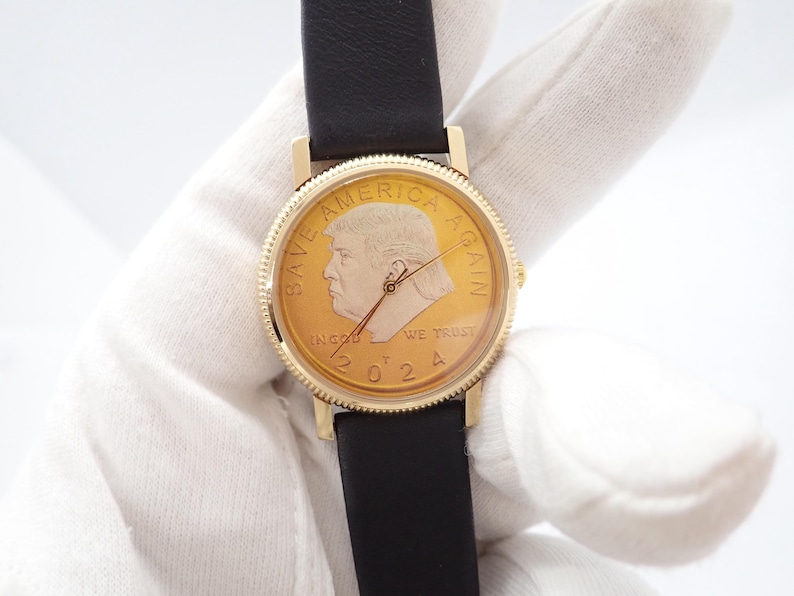 May include: Gold-toned wristwatch with a black leather band. The watch face features a profile portrait and the text "SAVE AMERICA AGAIN" and "IN GOD WE TRUST 2024". The watch has a gold-colored bezel.