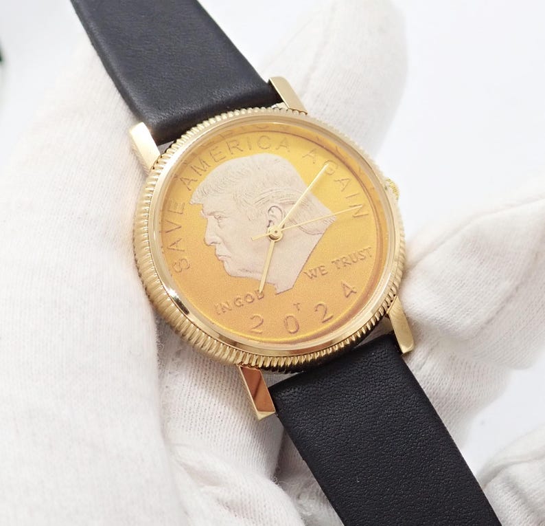 May include: Gold-toned wristwatch with a black leather band. The watch face features a gold coin design with a profile portrait, the text "SAVE AMERICA AGAIN", "IN GOD WE TRUST", and the year "2024".