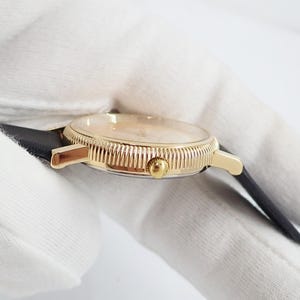 May include: A gold-toned wristwatch with a black leather band. The watch face is a light color, and the bezel has a textured design. The watch is a classic style.