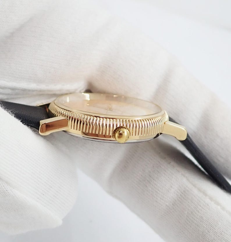 May include: A gold-toned wristwatch with a black leather band. The watch face is a light color, and the bezel has a textured design. The watch is a classic style.