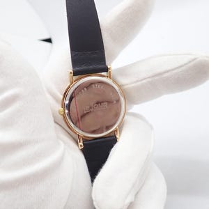 May include: A gold-toned wristwatch with a black leather band. The watch face is round with a gold bezel. The back of the watch is visible, displaying the text "LE JOUR" and "STAINLESS STEEL BACK".