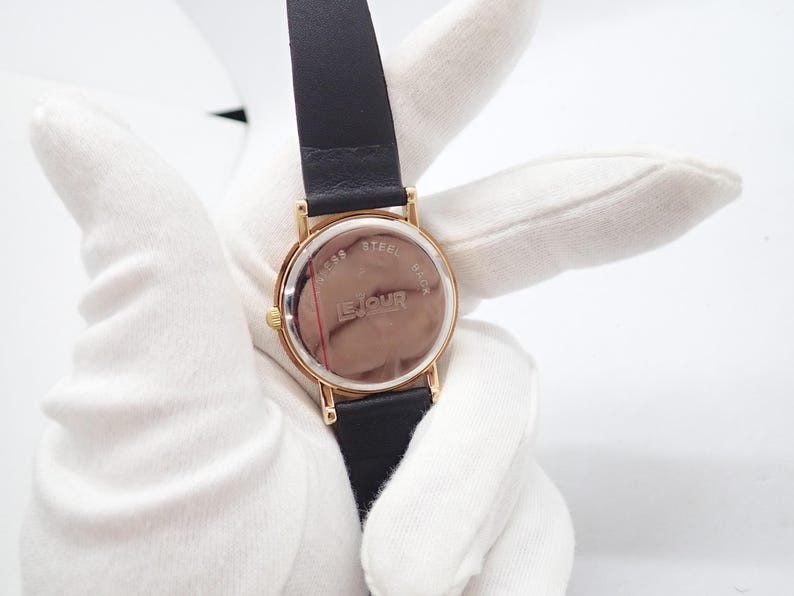 May include: A gold-toned wristwatch with a black leather band. The watch face is round with a gold bezel. The back of the watch is visible, displaying the text "LE JOUR" and "STAINLESS STEEL BACK".