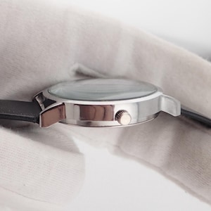 May include: A silver-toned wristwatch with a black leather band. The watch has a round face with a clear crystal and a small crown. The watch is shown from the side, with the band extending to the left and right.