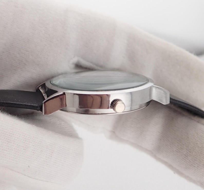 May include: A silver-toned wristwatch with a black leather band. The watch has a round face with a clear crystal and a small crown. The watch is shown from the side, with the band extending to the left and right.