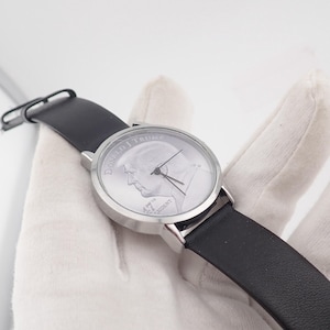 May include: A wristwatch with a silver-colored face featuring a profile image and the text "DONALD J. TRUMP 45TH PRESIDENT". The watch has a black leather band and a silver-colored case. The watch is being held by a gloved hand.