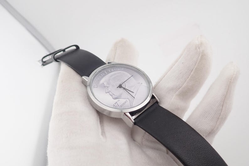 May include: A wristwatch with a silver-colored face featuring a profile image and the text "DONALD J. TRUMP 45TH PRESIDENT". The watch has a black leather band and a silver-colored case. The watch is being held by a gloved hand.
