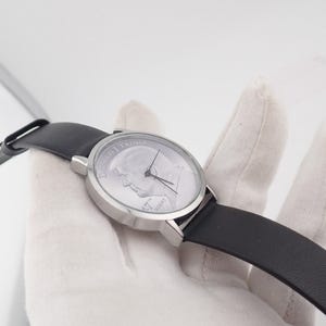 May include: A silver-toned wristwatch with a black leather band. The watch face features a portrait and text. The watch is held against a white background. The watch hands point to the time.