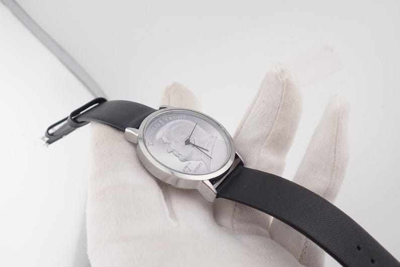 May include: A silver-toned wristwatch with a black leather band. The watch face features a portrait and text. The watch is held against a white background. The watch hands point to the time.