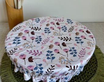 Op de afbeelding: Een ronde stoffen komafdekking met een elastische rand, voorzien van een witte achtergrond met een herhalend patroon van kleine roze, blauwe en bruine bloemen en bladeren. De hoes ligt over een kom, met keukengerei zichtbaar in de buurt.