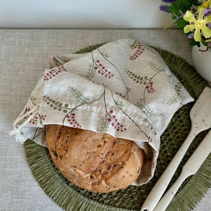 May include: A round loaf of bread is partially covered by a beige linen bread bag with a floral pattern in green and burgundy. The bread sits on a green placemat. Two white spatulas with colorful speckles are on the right.