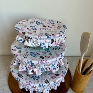 May include: A stack of three round fabric bowl covers with an elasticized ruffled edge. The white fabric features a repeating botanical pattern of small pink flowers, blue leaves, and dark brown fern-like sprigs across the surface of each cover.