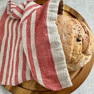 May include: A round loaf of crusty artisan bread with visible seeds sits on a circular wooden cutting board, partially covered by a folded beige and red striped fabric bag featuring a small white tag with black text on the upper left corner.