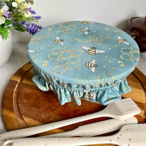 Op de afbeelding: Een turquoise stoffen komdeksel met een bijen- en honingraatpatroon, gerimpelde rand, en een set witte keukengerei met kleurrijke spikkels. De hoes ligt op een houten snijplank.