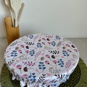 Op de afbeelding: Een witte taartovertrek met een bloemenpatroon met roze, blauwe en bruine botanische ontwerpen. De overtrek is over een rond object geplaatst, mogelijk een taart, en heeft een gerimpelde rand. Een houten keukengereihouder met witte kookgerei staat op de achtergrond.