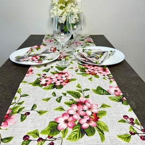 May include: A table setting featuring a floral table runner with pink blossoms and green leaves. White plates, napkins, and silverware are arranged on the runner. Two wine glasses and a vase of white flowers complete the elegant display.