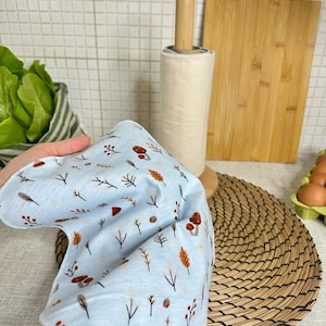 Op de afbeelding: Een lichtblauwe herbruikbare doek met een herhalend patroon van kleine bruine paddenstoelen, oranje bladeren en dunne zwarte takjes. De stof is zacht en flexibel, heeft afgeronde randen en wordt omhoog gehouden om de print te tonen.