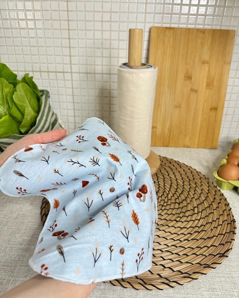 Op de afbeelding: Een lichtblauwe herbruikbare doek met een herhalend patroon van kleine bruine paddenstoelen, oranje bladeren en dunne zwarte takjes. De stof is zacht en flexibel, heeft afgeronde randen en wordt omhoog gehouden om de print te tonen.